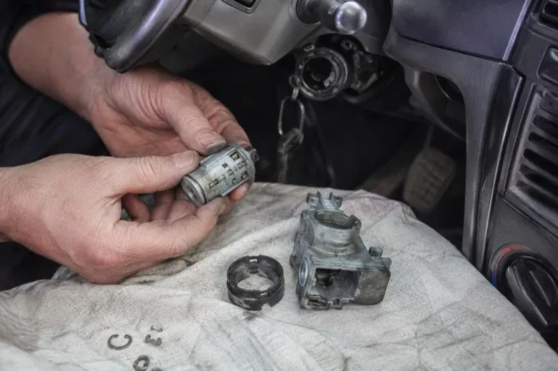 Hands holding a removed ignition cylinder beside a car steering column, with the housing and small parts on a cloth. — Ignition Lock Repair, Burlington