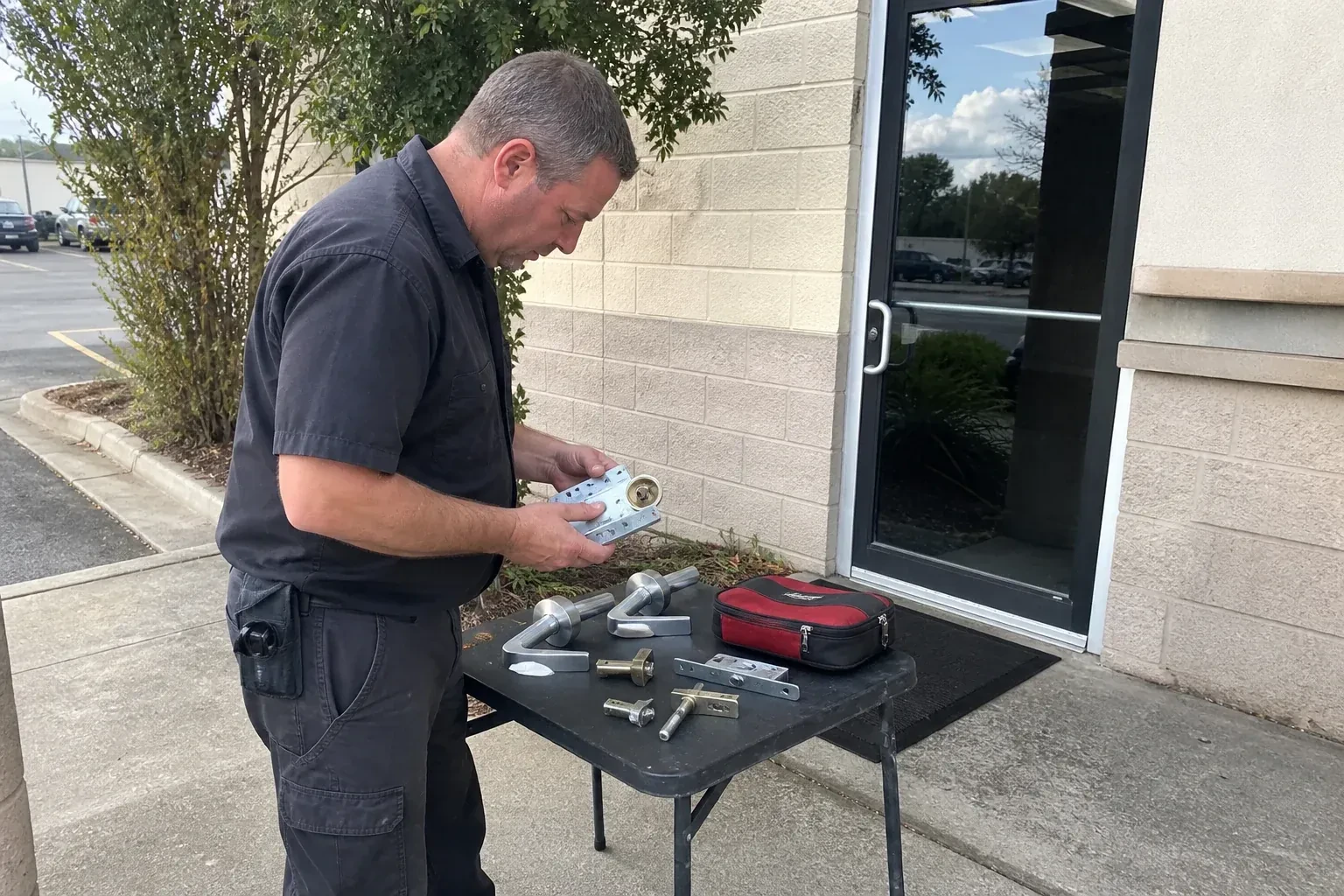 Technician examining detached commercial lock hardware on a table outside a shop entrance. — Business Lockout Service, Burlington